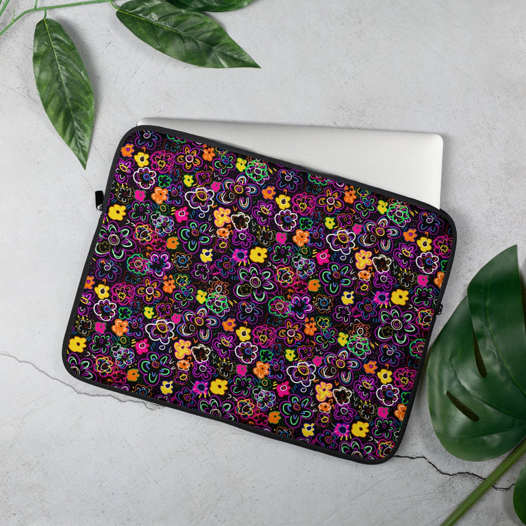 A laptop partially rests in the 15 inch 'In the Midnight Garden Laptop Sleeve' by My Favourite Colour is Rainbow.  The design features a vibrant floral design on a dark background.  It sits on a light grey surface surrounded by lush green leaves.