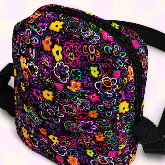 The In the Midnight Garden Mini Crossbody Bag by My Favourite Colour is Rainbow features a vibrant floral pattern with pink, orange, yellow, and green on a black background. It is made of canvas-type material, has internal and external pockets and an adjustable black strap with a buckle on one side that can be extended to crossbody length.