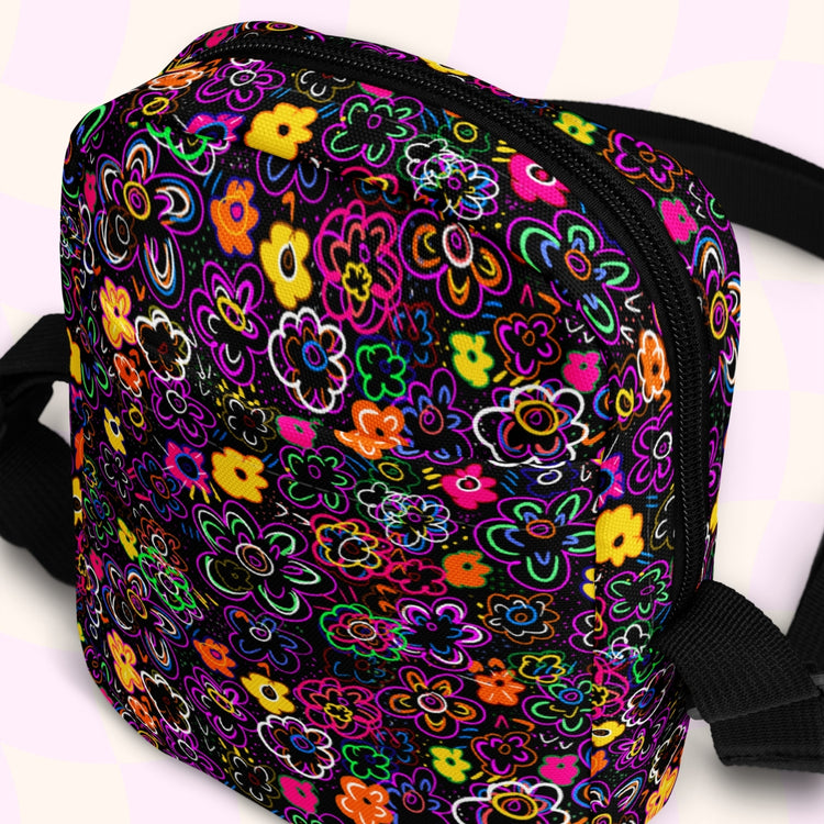 The In the Midnight Garden Mini Crossbody Bag by My Favourite Colour is Rainbow features a vibrant floral pattern with pink, orange, yellow, and green on a black background. It is made of canvas-type material, has internal and external pockets and an adjustable black strap with a buckle on one side that can be extended to crossbody length.