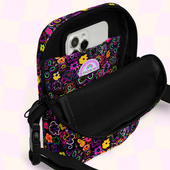 The In the Midnight Garden Mini Crossbody Bag by My Favourite Colour is Rainbow features a colourful floral print on a back background and an adjustable strap with a plastic buckle. Its open to show a smartphone placed in the inside pocket.