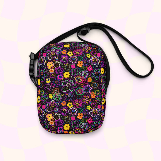 The In the Midnight Garden Mini Crossbody Bag by My Favourite Colour is Rainbow features a vibrant floral pattern with pink, orange, yellow, and green on a black background.  It is made of canvas-type material, has internal and external pockets and an adjustable black strap with a buckle on one side that can be extended to crossbody length.