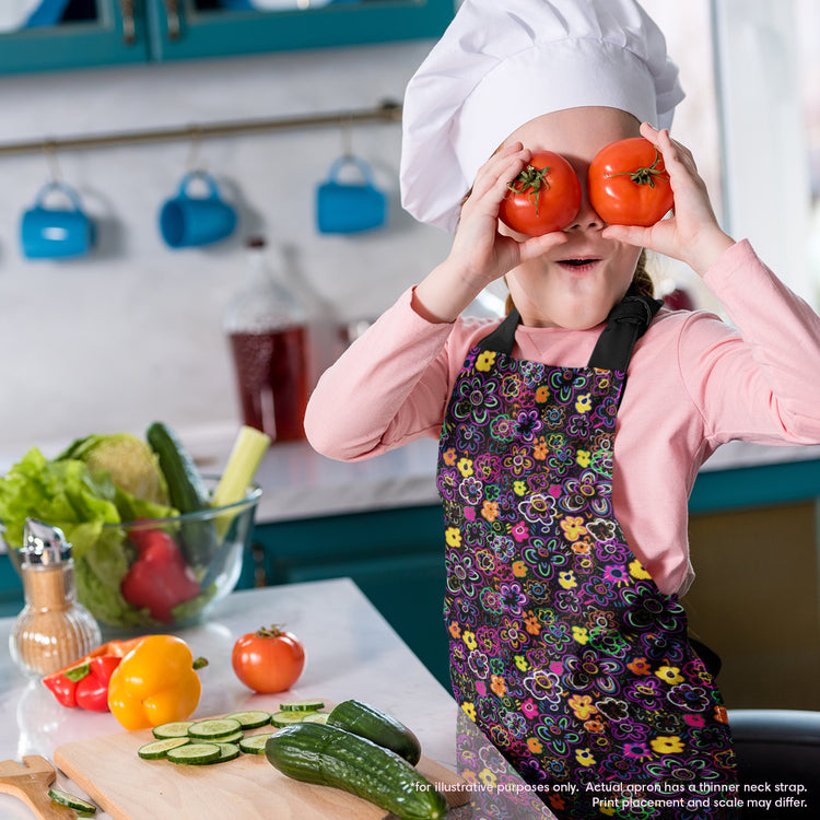 A child in a chefs hat and the In the Midnight Garden Apron by My Favourite Colour is Rainbow playfully places two tomatoes over their eyes in a lively kitchen. Cucumbers and bell peppers line the counter, surrounded by utensils and a pitcher, enhancing the vibrant cooking experience.  The apron features a striking design of multicoloured flowers on a black background.