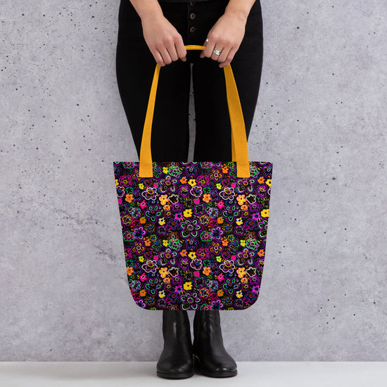 A person holds the In the Midnight Garden Tote Bag from My Favourite Colour is Rainbow, featuring a vibrant floral pattern. They wear black pants and boots, while bright yellow handles of the bag add a pop of colour to their outfit.