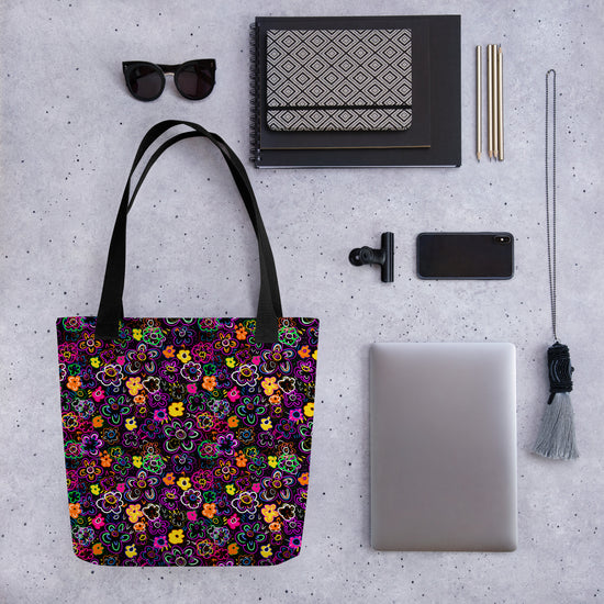 Flat lay of items on a gray surface, featuring the In the Midnight Garden Tote Bag by My Favourite Colour is Rainbow, sunglasses, notebooks, pens, a smartphone, a clip, a closed laptop, and a tassel accessory. The bag features colourful hand-drawn flowers on a black background.
