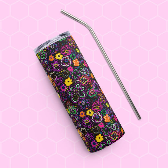 The In the Midnight Garden Stainless Steel Tumbler by My Favourite Colour is Rainbow, featuring a colorful floral pattern on a black background, rests diagonally on a pink hexagonal-patterned surface with a sleek metal straw laid parallel for an elegant touch.