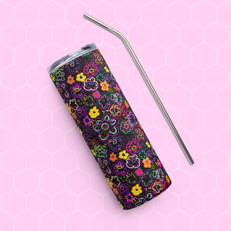 The In the Midnight Garden Stainless Steel Tumbler by My Favourite Colour is Rainbow, featuring a colorful floral pattern on a black background, rests diagonally on a pink hexagonal-patterned surface with a sleek metal straw laid parallel for an elegant touch.