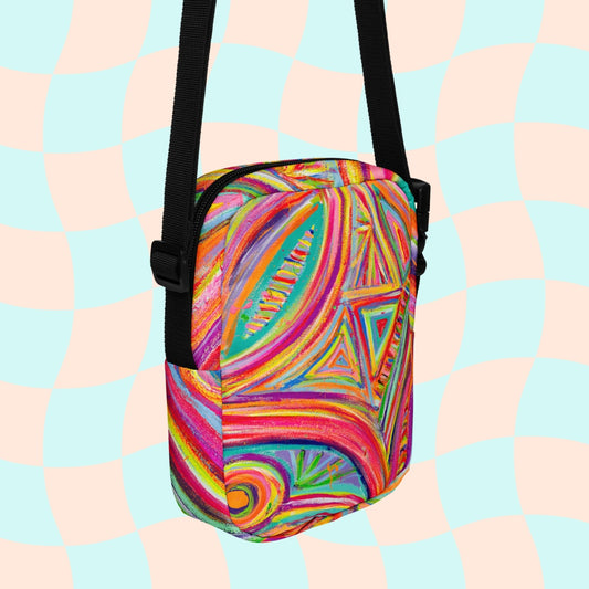 Discover the Karma Kaleidoscope Mini Crossbody Bag by My Favourite Colour is Rainbow, featuring a lively pattern of bold pink, orange, blue, and green swirls and geometric shapes. It is made of canvas-type material, has internal and external pockets and an adjustable black strap with a buckle on one side that can be extended to crossbody length.