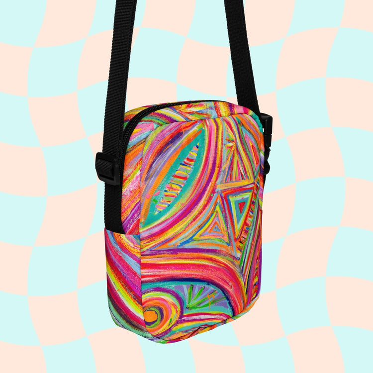 Discover the Karma Kaleidoscope Mini Crossbody Bag by My Favourite Colour is Rainbow, featuring a lively pattern of bold pink, orange, blue, and green swirls and geometric shapes. It is made of canvas-type material, has internal and external pockets and an adjustable black strap with a buckle on one side that can be extended to crossbody length.