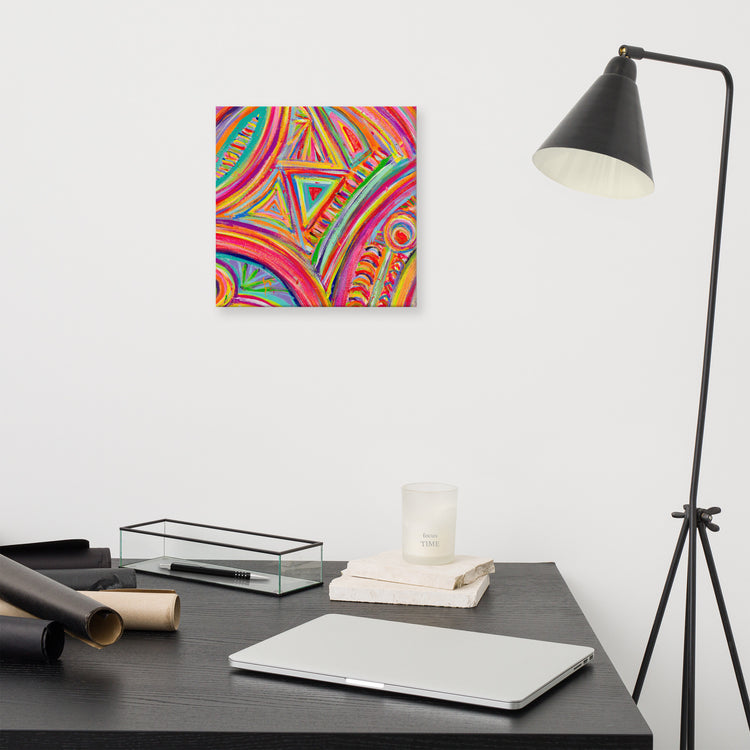A sleek, modern desk features a closed laptop, rolled papers, and a candle beneath a black floor lamp. Hanging on the white wall is the vibrant Karma Kaleidoscope Canvas Artwork by My Favourite Colour is Rainbow, showcasing circular and triangular patterns.