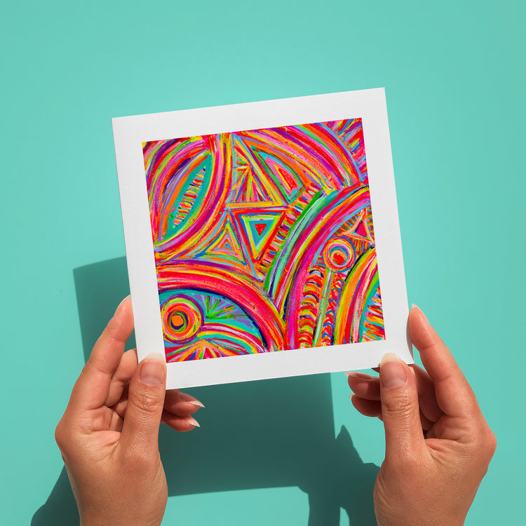 Two hands hold a vibrant Karma Kaleidoscope Greeting Card by My Store against a teal background, showcasing bold patterns of swirling red, orange, green, and blue with dynamic geometric shapes like circles and triangles.