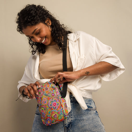 A curly-haired woman smiles as she adjusts her Love in Neon Mini Crossbody Bag by My Favourite Colour is Rainbow. This colorful bag, with adjustable straps and water resistance, complements her tan top, white shirt, and blue jeans against a neutral background.