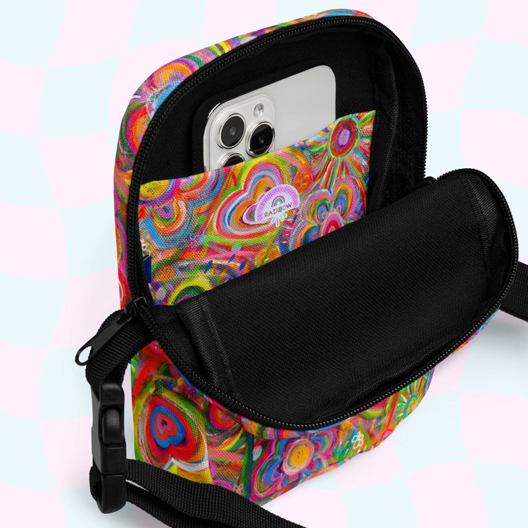 The Love in Neon Mini Crossbody Bag by My Favourite Colour is Rainbow, featuring a colorful psychedelic pattern and black adjustable strap, is open with a smartphone peeking out. The pastel teal and pink checkerboard background adds to the vibrant look.