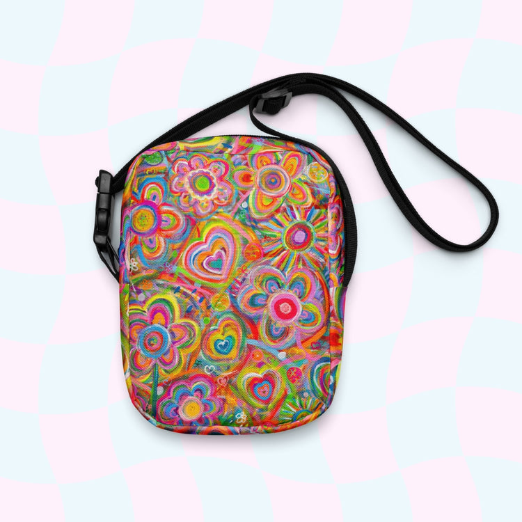 The Love in Neon Mini Crossbody Bag by My Favourite Colour is Rainbow features vibrant, psychedelic floral and heart patterns in pink, yellow, green, and blue. It includes a black adjustable strap on a pastel checkerboard background and is water-resistant.