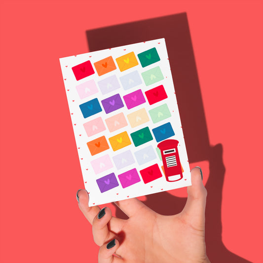 A hand holds My Stores Love Letter Valentines card, featuring a vibrant grid of envelopes with hearts on a red background and a charming red postbox icon in the bottom right corner, radiating rainbow joy.