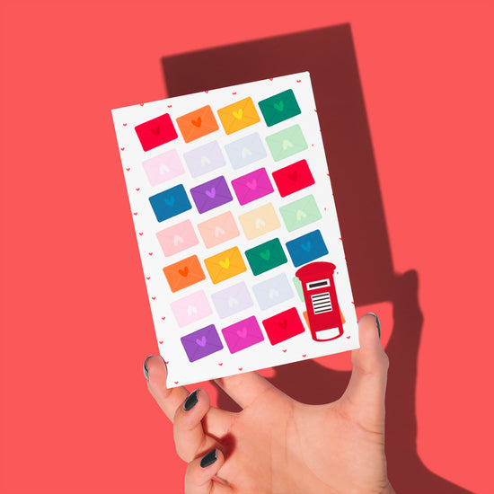 A hand holds My Stores Love Letter Valentines card, featuring a vibrant grid of envelopes with hearts on a red background and a charming red postbox icon in the bottom right corner, radiating rainbow joy.