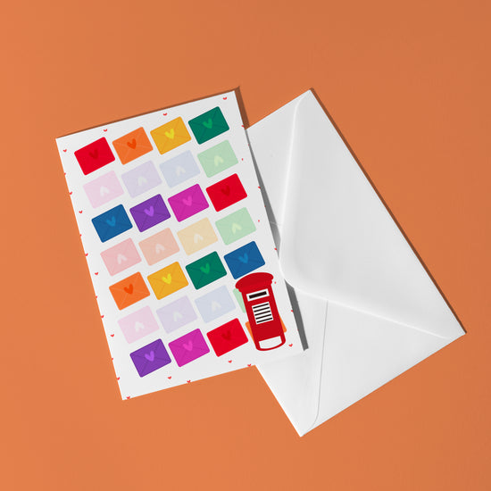 The Love Letter Valentines Card by My Store showcases 23 colorful envelope illustrations and a red postbox, creating a vibrant display of rainbow joy against an orange background. It comes with a white envelope.