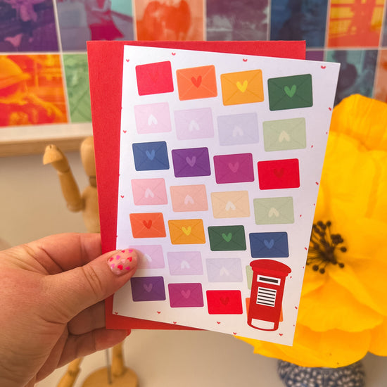 A hand with pink heart nail art holds My Stores Love Letter Valentines Card, featuring colorful envelopes and a charming red postbox. A bright yellow flower enhances the allure against a blurred, rainbow-filled backdrop.