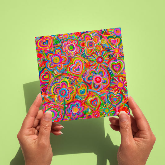 Two hands hold a vibrant square artwork from My Stores Love in Neon Greeting Card collection, showcasing glowing patterns of overlapping hearts and spirals in radiant colors like red, pink, orange, green, and blue. A light green background enhances the vividness.
