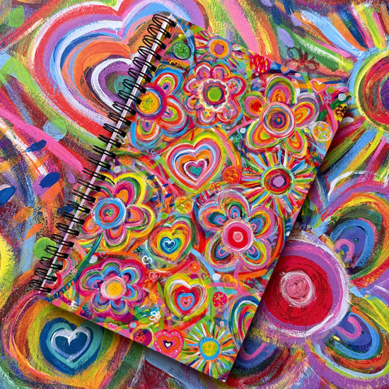 The Love in Neon Notebook from My Store, featuring a spiral binding and decorated with vibrant swirling patterns of hearts and flowers in red, pink, yellow, and blue, lies on a lively abstract background—an ideal addition to any creativity-driven stationery collection.