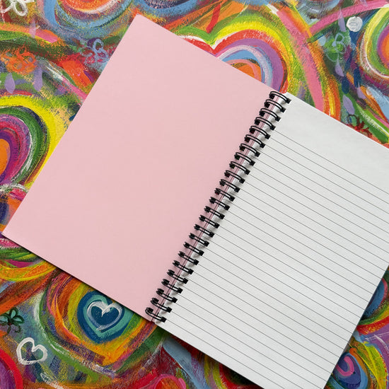 The Love in Neon Notebook from My Store rests on a vibrant surface with its left page blank and pink, while the right page features lined white paper. The colorful background with swirling hearts and bright abstract designs inspires creativity.