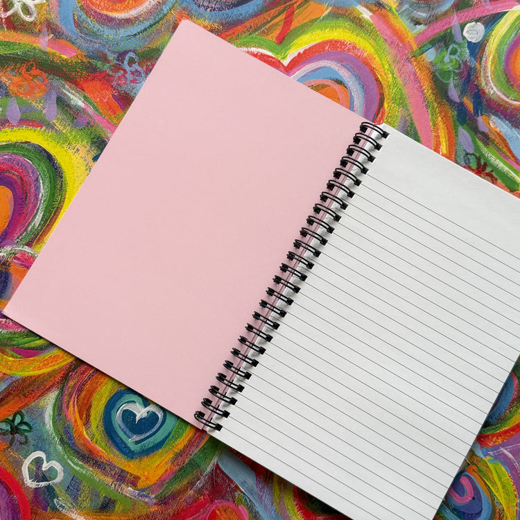 The Love in Neon Notebook from My Store rests on a vibrant surface with its left page blank and pink, while the right page features lined white paper. The colorful background with swirling hearts and bright abstract designs inspires creativity.