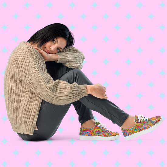 A woman sits with her head on her knee in a beige sweater and dark pants, wearing My Favourite Colour is Rainbows Love in Neon canvas sneakers featuring a colorful print for vibrant style against a pink background with light blue star patterns.