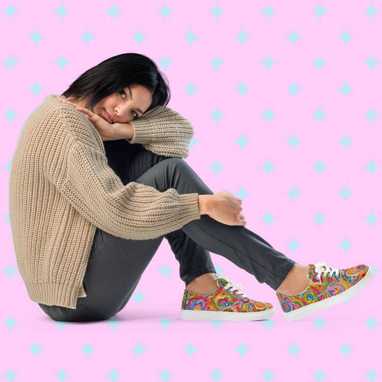 A woman sits with her head on her knee in a beige sweater and dark pants, wearing My Favourite Colour is Rainbows Love in Neon canvas sneakers featuring a colorful print for vibrant style against a pink background with light blue star patterns.