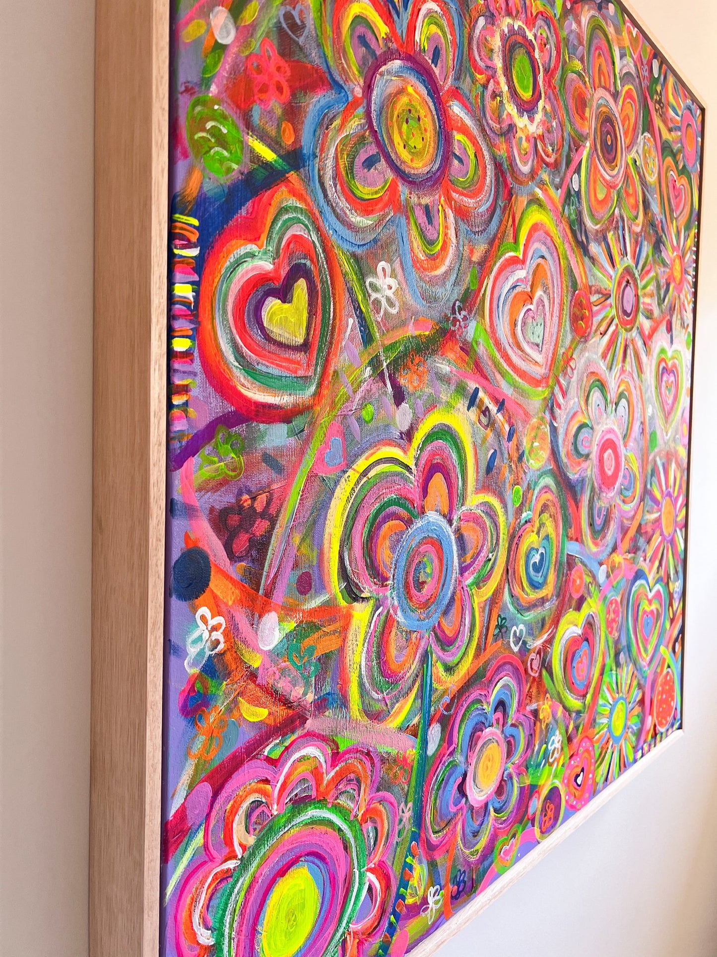 The Love In Neon Framed Original Painting by My Store is a colorful abstract with vibrant flowers and heart shapes in pink, green, blue, and neon tones. This acrylic artwork covers the entire surface and is elegantly set within a wooden frame.