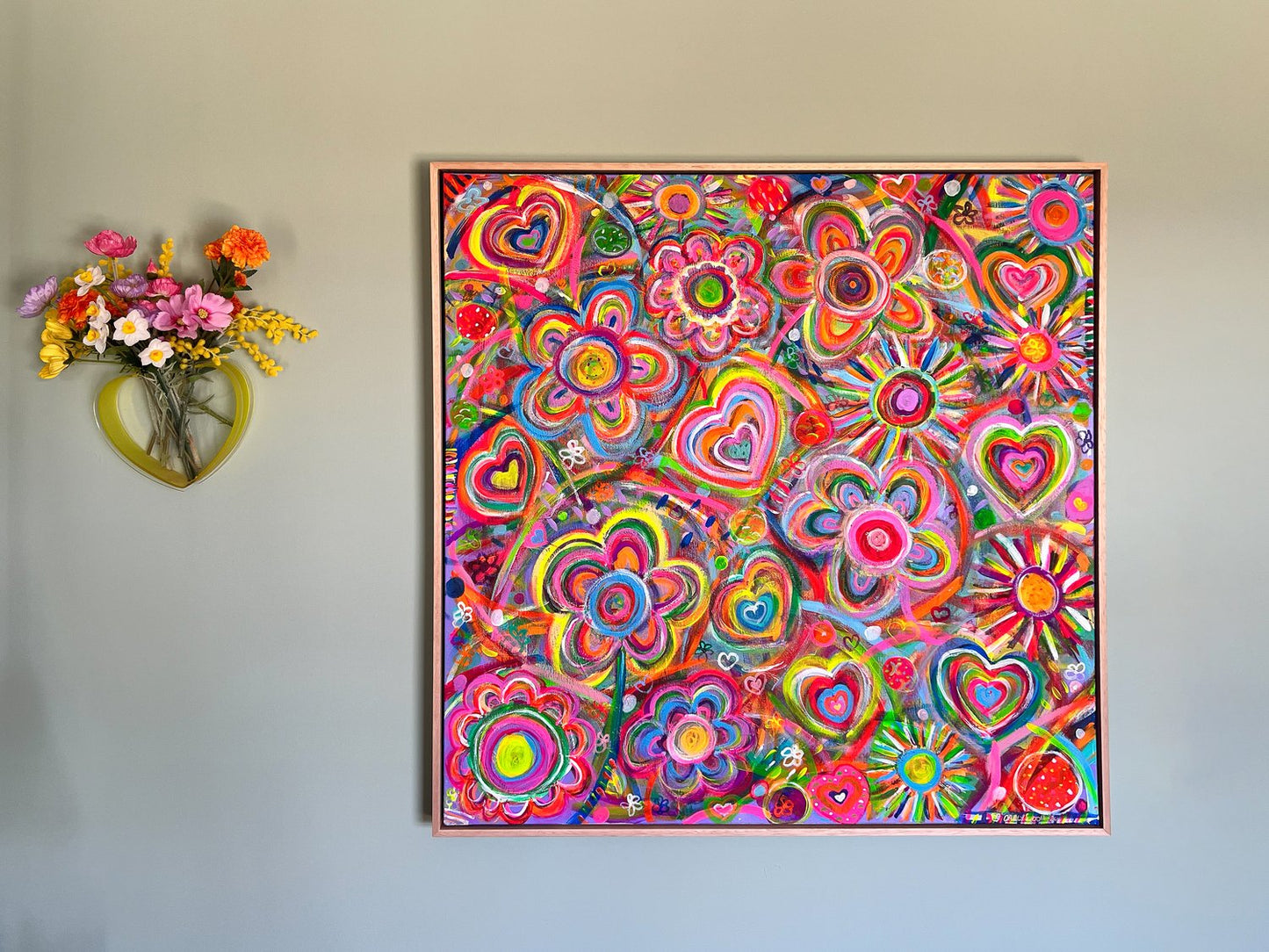 The Love In Neon framed original painting by My Store, featuring vibrant acrylic heart and flower patterns, is displayed on a light gray wall. To its left, a heart-shaped vase contains a bouquet of bright neon-toned flowers, enhancing the artworks vivid allure.