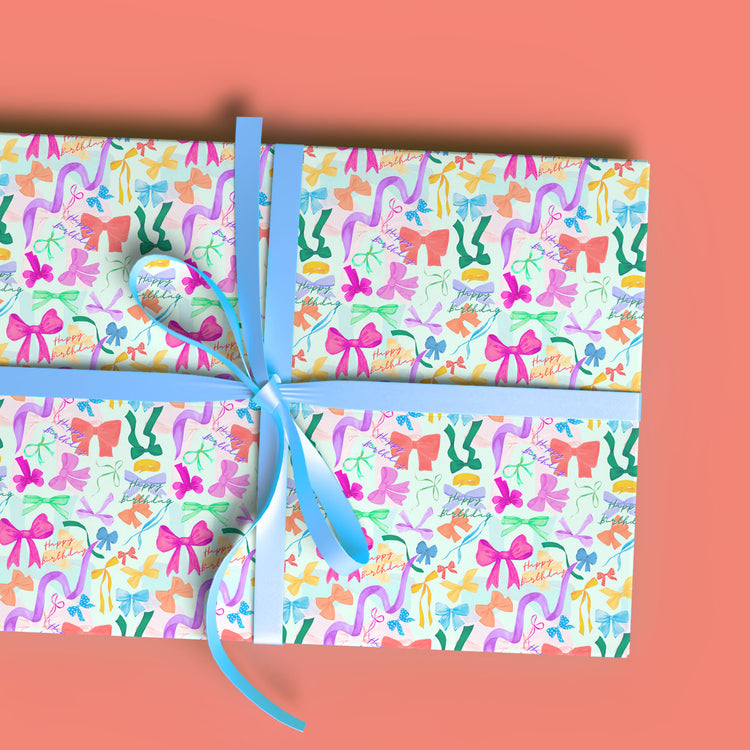 A gift wrapped in My Favourite Colour is Rainbows Multicolour Bow Birthday Wrapping Paper, featuring vibrant patterns of bows, shoes, and hats in pink, blue, orange, and green. It is tied with a light blue ribbon and shown against a coral background.