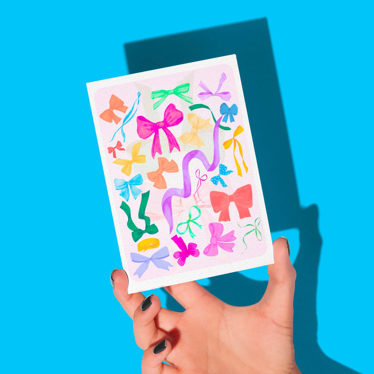 A hand with painted nails holds the My Store Multicolour Bow Greeting Card against a bright blue background, showcasing various colorful bows that cast distinct shadows.