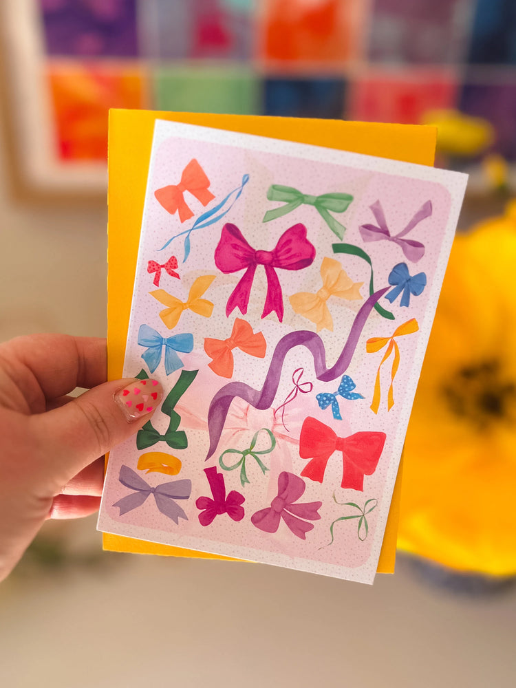 A hand holds My Stores Multicolour Bow Greeting Card, adorned with vibrant bow illustrations. In the blurred background blooms a bright yellow flower. Their nails, painted pink with white polka dots, add extra charm to this stylish scene.