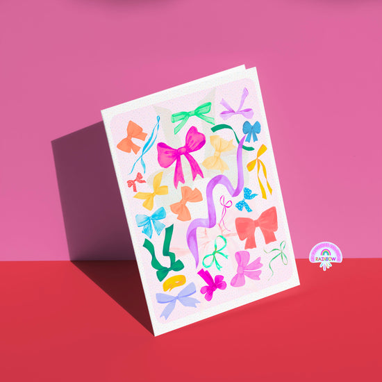 The Multicolour Bow Greeting Card by My Store stands against a pink background, adorned with vibrant bows and ribbons. A small rainbow logo with Rainbow at the bottom right adds an extra touch to this stylish, versatile birthday card.