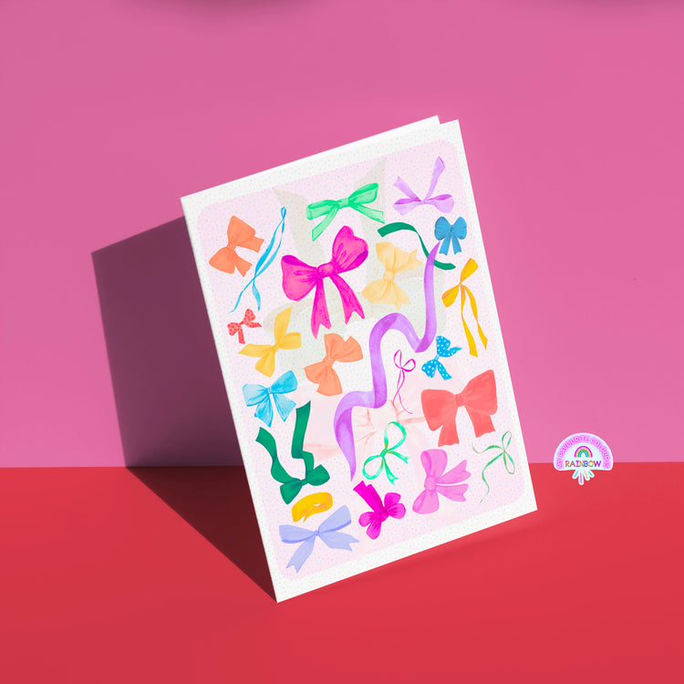 The Multicolour Bow Greeting Card by My Store stands against a pink background, adorned with vibrant bows and ribbons. A small rainbow logo with Rainbow at the bottom right adds an extra touch to this stylish, versatile birthday card.