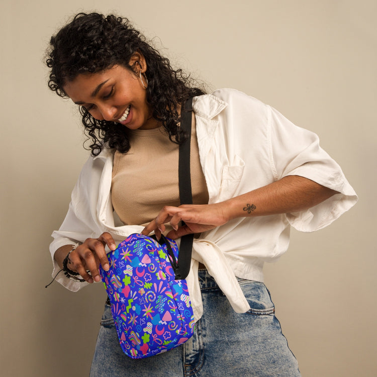 Wearing a white shirt over a beige top and jeans, a person with curly hair smiles while admiring their Neon Carnival Mini Crossbody Bag by My Favourite Colour is Rainbow. The bag has adjustable straps and stands out against the plain beige background.