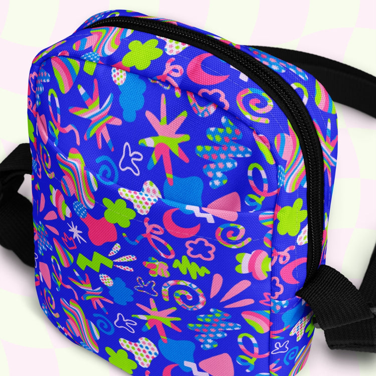 The Neon Carnival Mini Crossbody Bag by My Favourite Colour is Rainbow showcases a vibrant blue background with abstract patterns: stars, swirls, and shapes in pink, green, and yellow. Black zipper and adjustable straps complement the neon design on a light checkered backdrop.