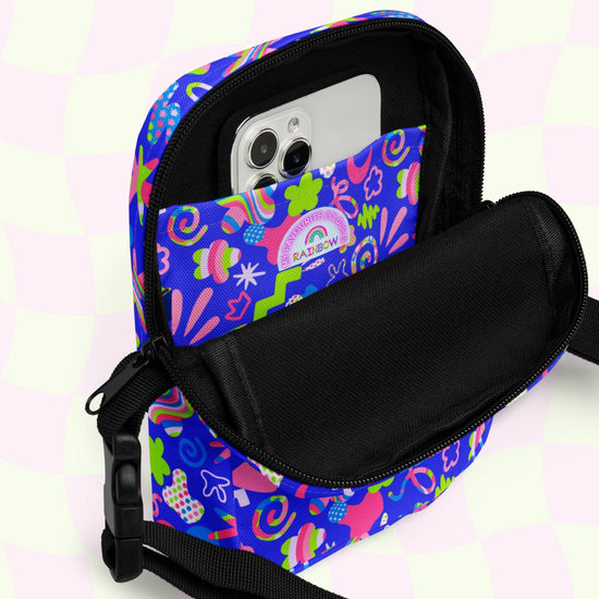 A Neon Carnival Mini Crossbody Bag by My Favourite Colour is Rainbow showcases a smartphone inside its pocket, with blue, pink, green, and white colors. The adjustable straps promise a perfect fit. It sits against a pastel checkered pink and green background.