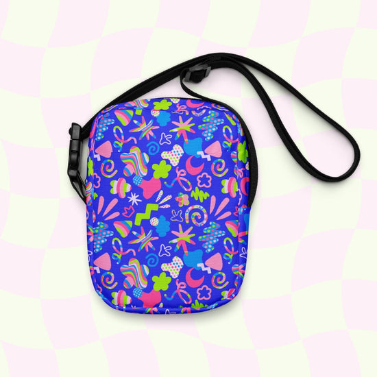 The Neon Carnival Mini Crossbody Bag by My Favourite Colour is Rainbow features a vibrant print of colorful abstract shapes, including stars, swirls, and dots on bright blue with a pastel pink and green checkered pattern backdrop. The black adjustable strap adds the perfect complement.