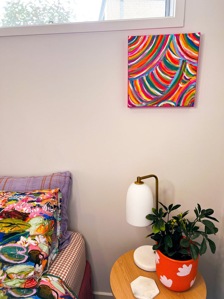 A cozy room corner showcases a bed with vibrant bedding, a small wooden table holding a plant and lamp, and the Neon Candy Floss Original Painting from My Store. This colorful acrylic on canvas art complements the natural light streaming through the window above.