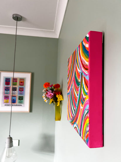 The Neon Candy Floss Original Painting by My Store, an acrylic on canvas with rainbow swirls, hangs on a light green wall. Below, a vase with vibrant flowers sits beside a framed print. A pendant light from the ceiling illuminates the scene like the painting itself.