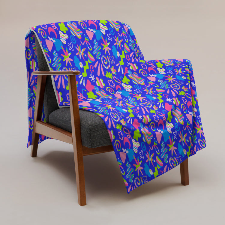 A wooden armchair with a dark cushion features the vibrant Neon Carnival Throw Blanket by My Favourite Colour is Rainbow, adorned with pink hearts, green stars, and yellow spirals. The cosy fabric adds warmth against a neutral background.