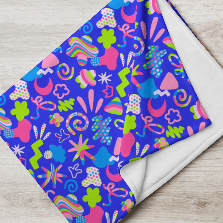 The Neon Carnival Throw Blanket by My Favourite Colour is Rainbow features a lively design with pink, green, and orange spirals, zigzags, and flowers on one side, with plain white on the reverse. Its elegantly laid on a light wooden surface.