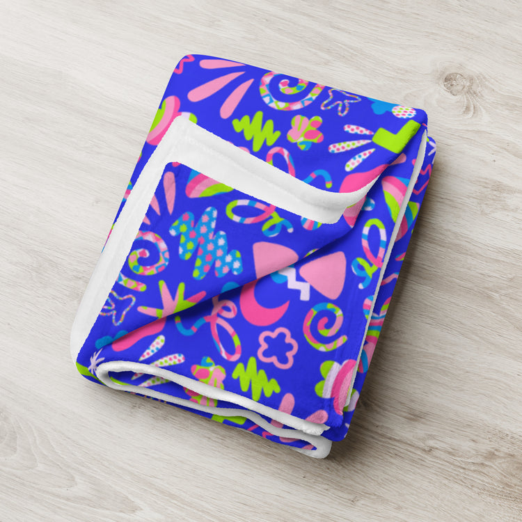 The Neon Carnival Throw Blanket by My Favourite Colour is Rainbow features a vibrant design of abstract shapes like leaves, flowers, and swirls in pink, green, and yellow on a bright blue background. Its displayed neatly folded on light wood to highlight its colorfulness and coziness.