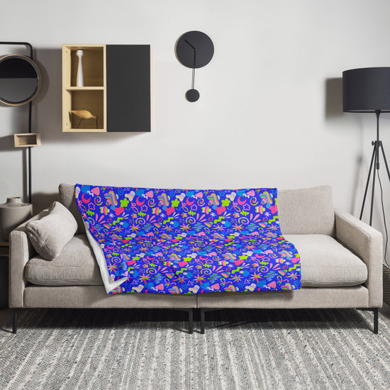 A beige couch is in a minimalist living room, highlighted by a Neon Carnival Throw Blanket from My Favourite Colour is Rainbow. A black shelf with decor and a round clock adorns the wall, while a tall black floor lamp enhances the cozy ambiance on the right.