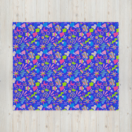 The Neon Carnival Throw Blanket by My Favourite Colour is Rainbow showcases a vibrant abstract pattern in pink, green, and purple with flowers, leaves, and swirls. Made from cosy fabric, its displayed flat on a light wood background.