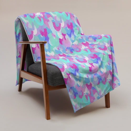 A wooden armchair with a gray cushion is draped with the Pastel Rainbow Throw Blanket by My Favourite Colour is Rainbow, featuring vibrant abstract shapes in pink, purple, blue, and green hues. The cozy scene is set against a neutral beige background.