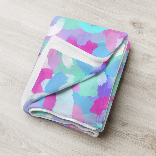 A folded Pastel Rainbow Throw Blanket by My Favourite Colour is Rainbow features a vibrant abstract design in pink, blue, green, and purple and rests invitingly on a light wooden surface.