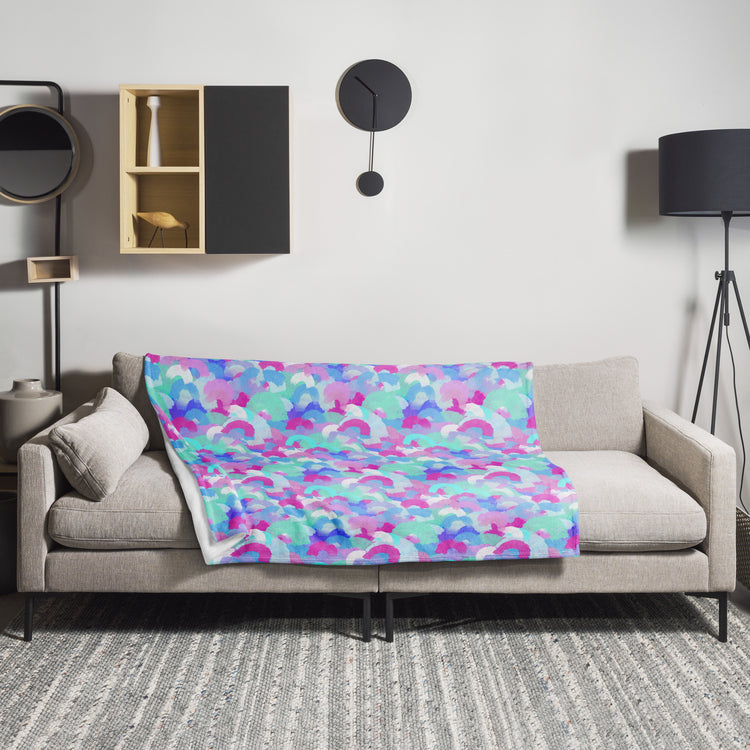 A modern living room includes a light gray sofa with a cozy Pastel Rainbow Throw Blanket by My Favourite Colour is Rainbow, featuring purple, blue, and pink patterns. Above, a minimalist black clock pairs with a black floor lamp and shelf showcasing vibrant decor items.
