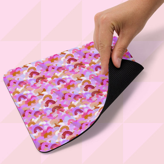 A hand holds the Pink Rainbow Mouse Pad from My Favourite Colour is Rainbow, featuring overlapping pink, purple, and orange semicircles on a black base. The backdrop showcases a light pink geometric pattern.