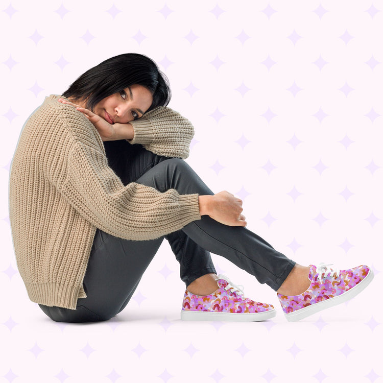 A woman sits on the floor wearing a beige knit sweater, dark pants, and Pink Rainbow Canvas Sneakers by My Favourite Colour is Rainbow. She rests her chin on her knee in contemplation against a backdrop with light purple star patterns.
